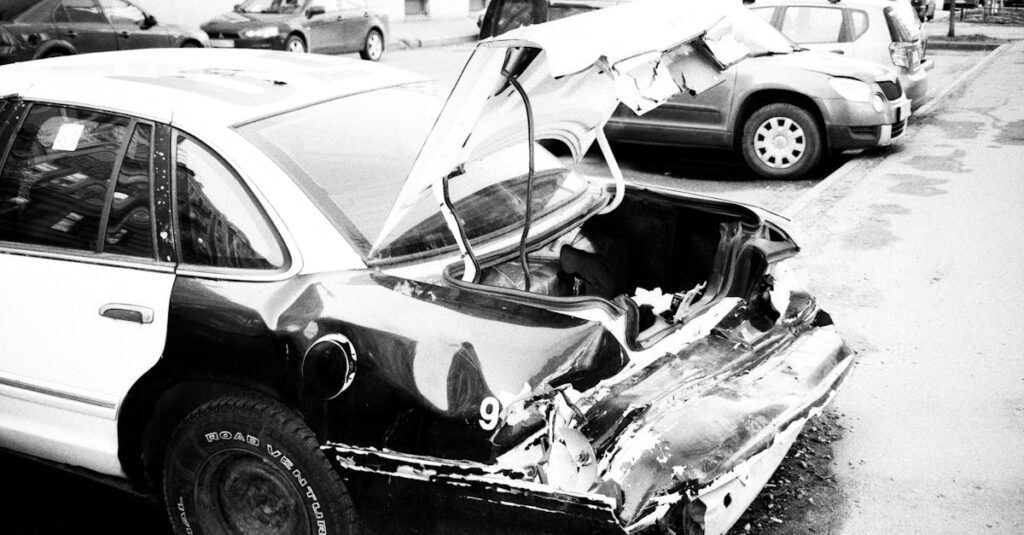 A black and white photo of a wrecked car on an urban street, highlighting vehicle damage.