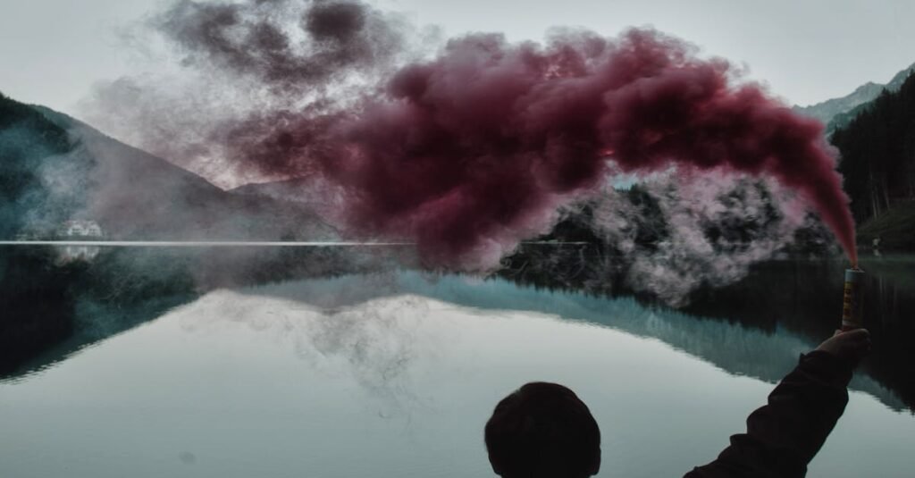 Silhouette holding smoke bomb by a tranquil lakeside with mountain reflection.