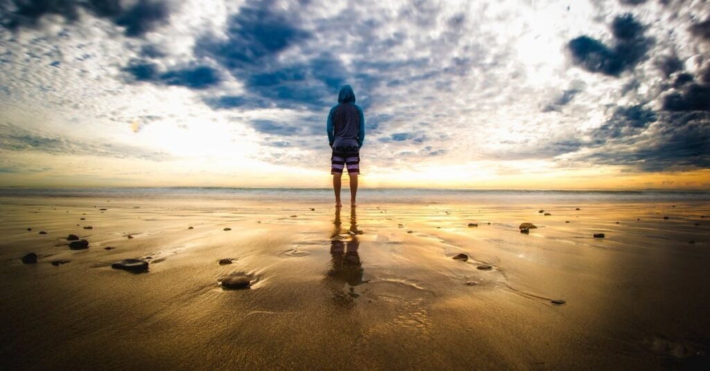 A lone figure stands on a tranquil beach during a vivid sunset, reflecting on the wet sand.