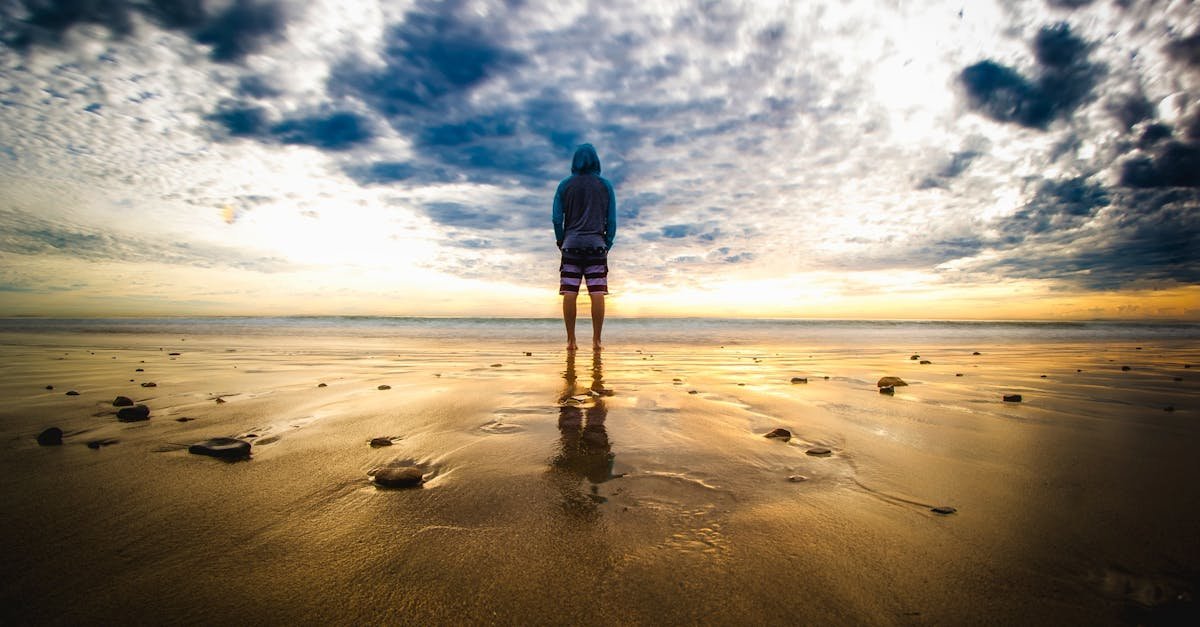 A lone figure stands on a tranquil beach during a vivid sunset, reflecting on the wet sand.