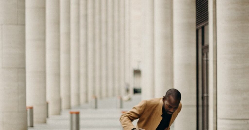 A black man in a fashionable coat walks through an architectural columned pathway.
