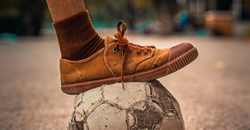 An aged soccer ball under a foot in casual footwear on an outdoor surface.