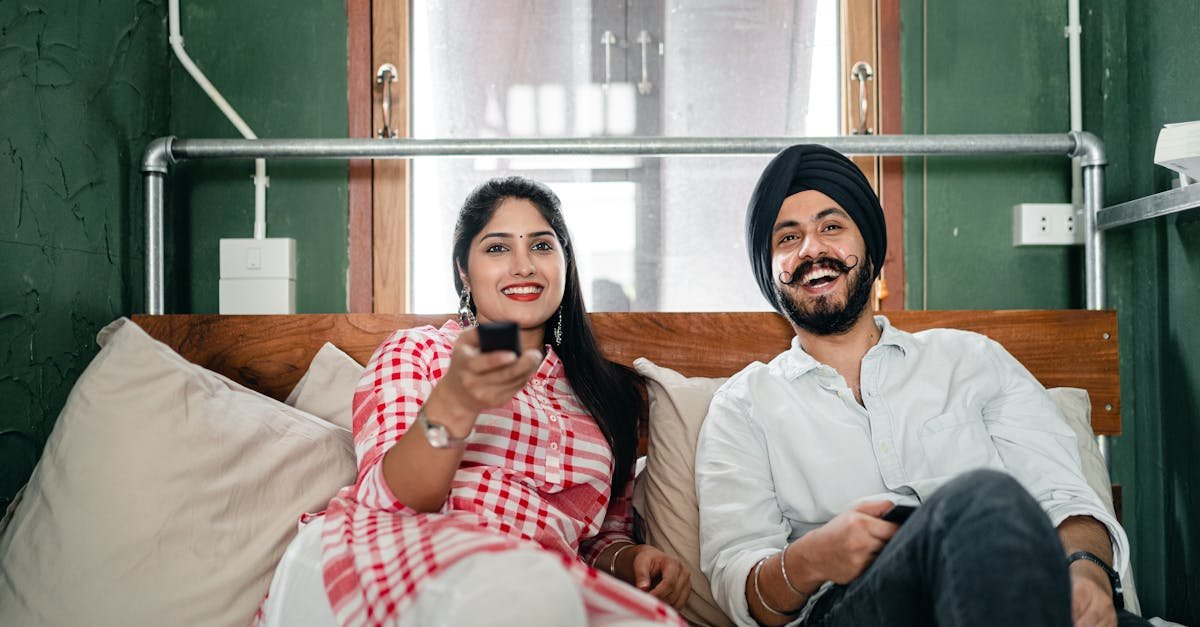 Cheerful wife with bindi on forehead wearing plaid tunic with white trousers using TV remote control for channel switching while lying on bed with laughing Sikh husband in turban with stylish beard and twisted mustache