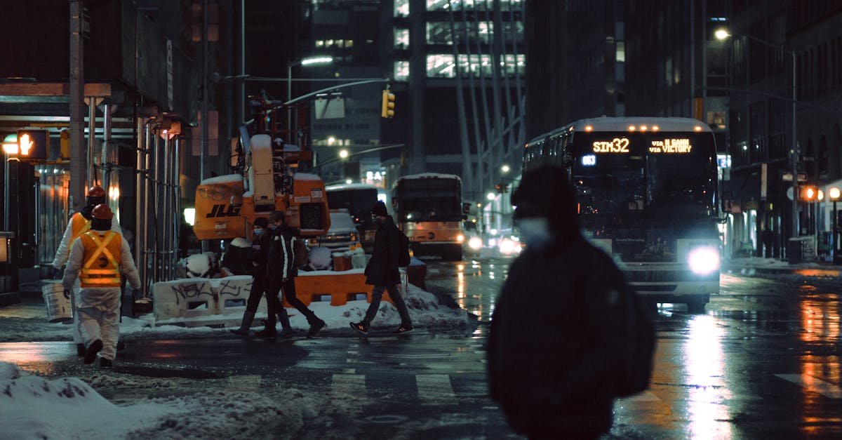 People in medical masks strolling on crosswalk near roadway with glowing buses on dark street during winter evening in city