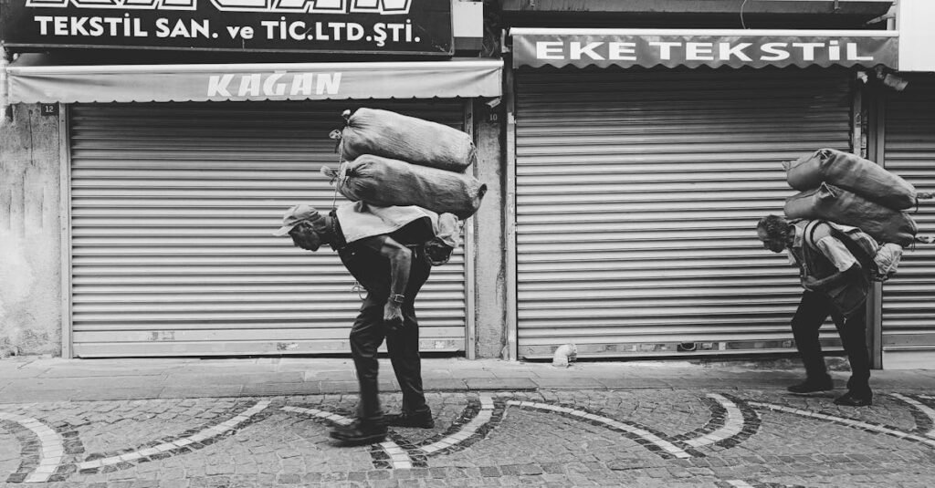 Black and white photo of two elderly men carrying heavy sacks on an urban street with closed shops.
