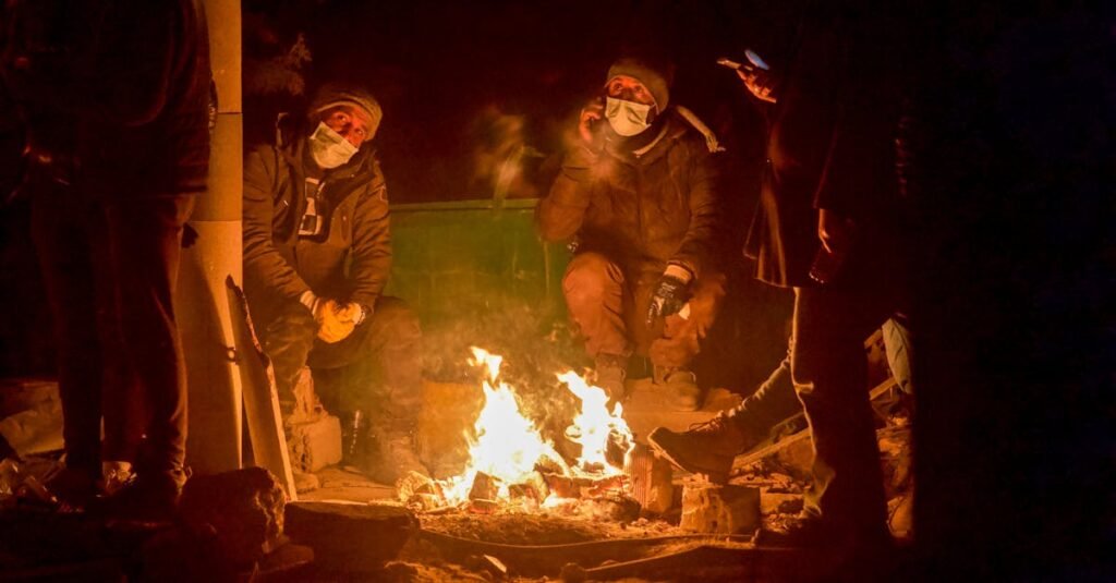 A group of individuals sitting around a campfire at night in Kahramanmaraş, Türkiye, wearing warm clothing and masks.