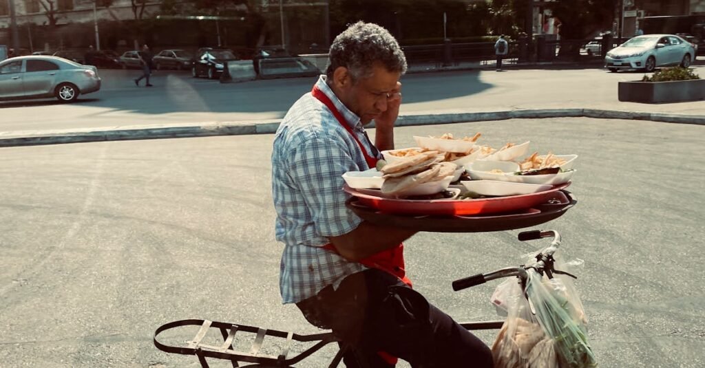 A man skillfully balances food trays on a bicycle in an urban Cairo street, showcasing daily life in the city.