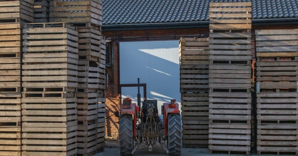 Red tractor positioned among towering stacks of wooden pallets outside a warehouse.