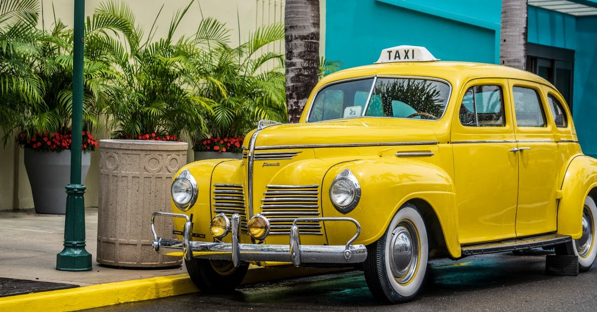 Vintage yellow taxi car parked on an urban street with lush green plants and palm trees.