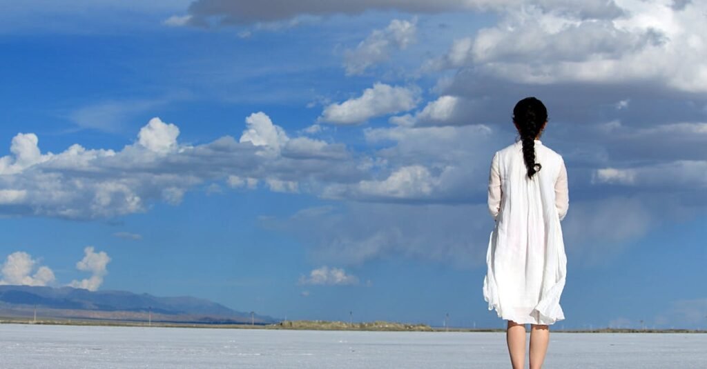 Woman stands alone on a vast salt flat under a bright blue sky with clouds.