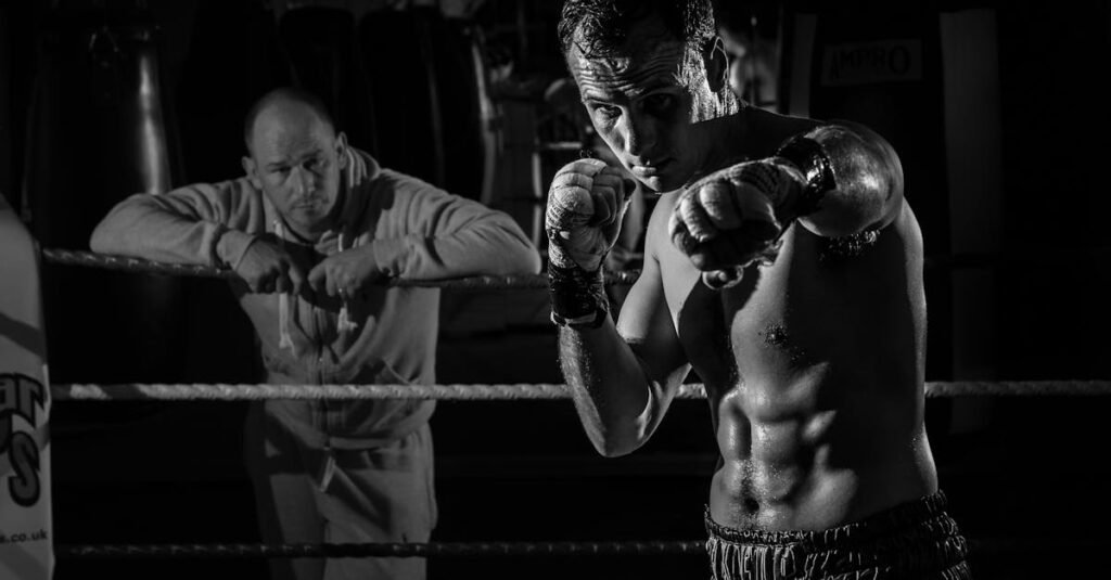 Boxer practicing in the ring with coach observing, captured in dramatic lighting.