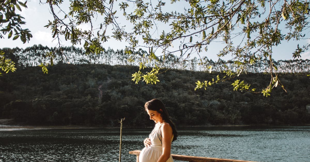 A pregnant woman stands gracefully on a dock by a tranquil lake, surrounded by nature.