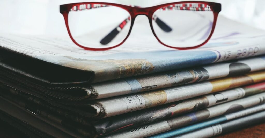 Red eyeglasses resting on a stack of newspapers symbolize reading and information.