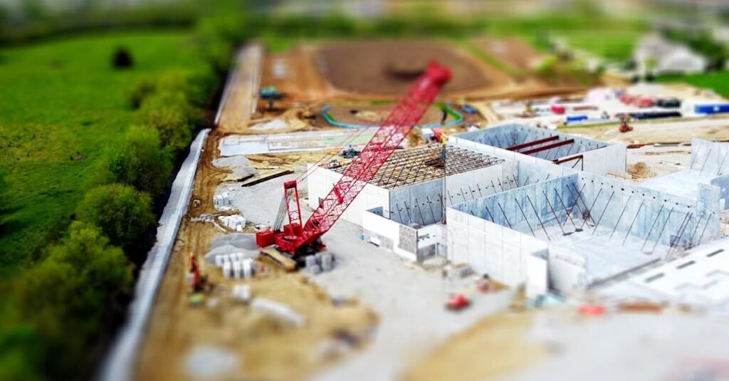Aerial view of a construction site with a red crane amidst green fields and concrete buildings.