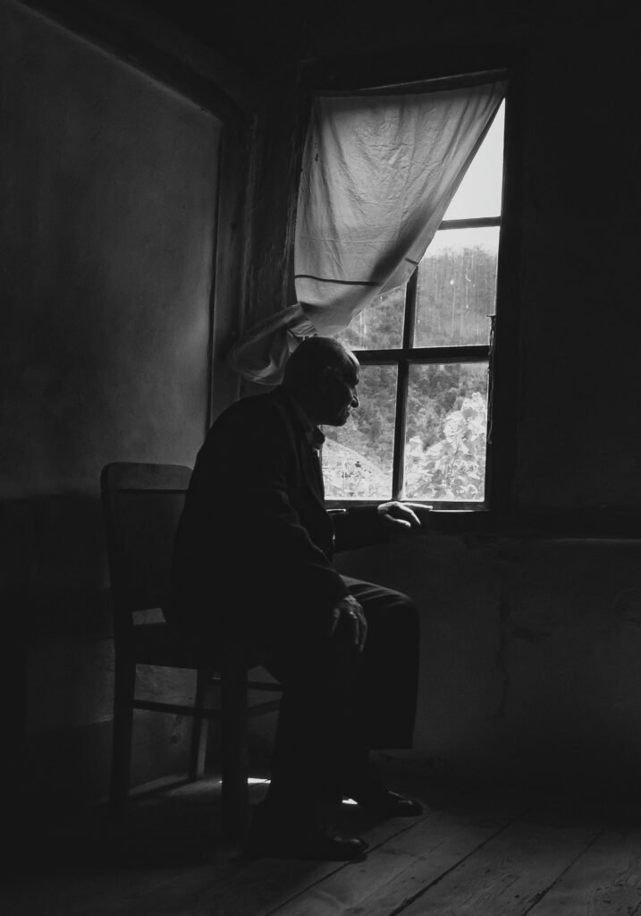 A black and white image of an elderly man sitting alone by a window, evoking loneliness and contemplation.
