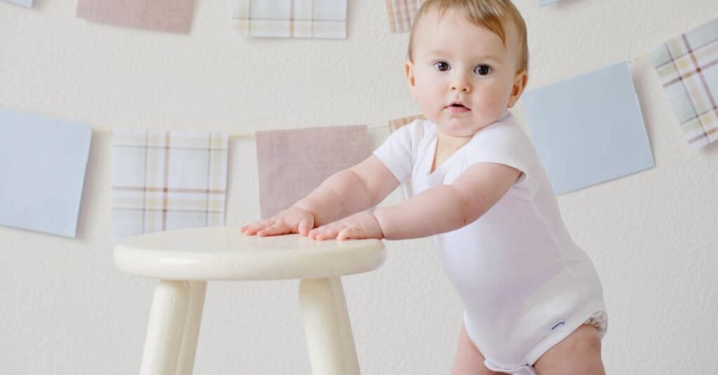 Cute baby standing by a stool indoors with a playful background.