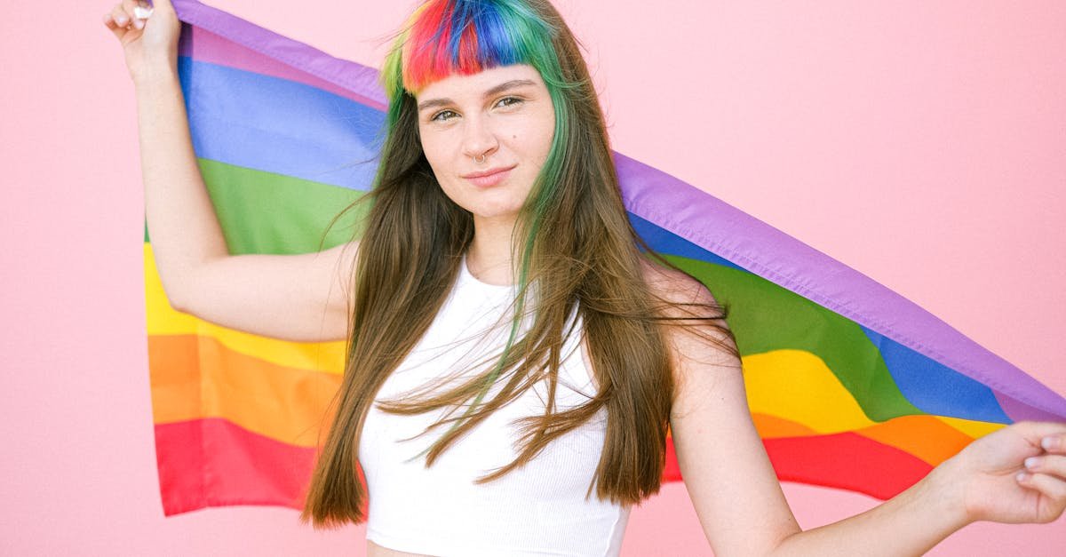A woman with rainbow bangs holding a pride flag against a pink background, symbolizing LGBTQ pride.