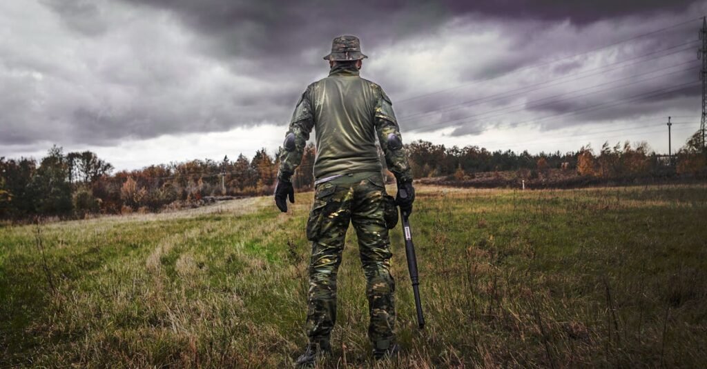 A soldier in camouflage uniform with a rifle stands in a grassy field under a cloudy sky.