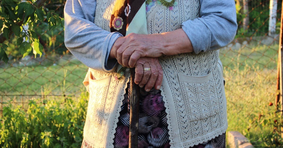 Elderly woman in garden holding a walking stick, wearing a knitted vest.