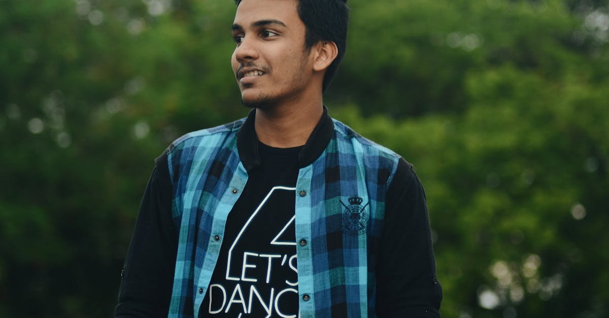 A young man in a casual plaid shirt smiling while standing outside against greenery.