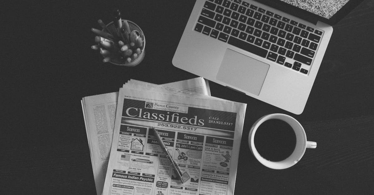 A black and white image of a laptop, newspaper, and coffee cup on a desk.