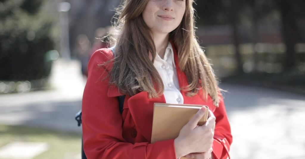 Young woman in a red coat smiling while holding a book outside on a sunny day.