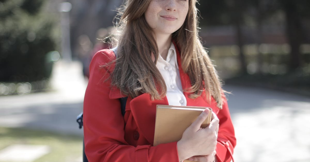 Young woman in a red coat smiling while holding a book outside on a sunny day.