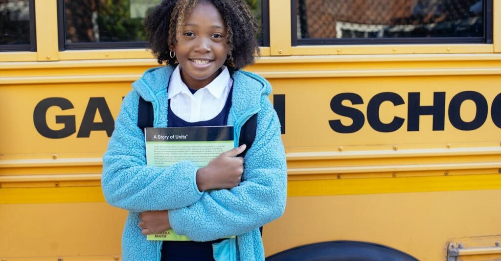 Cheerful African American girl in school uniform and warm jacket standing with textbook near yellow school bus and looking at camera