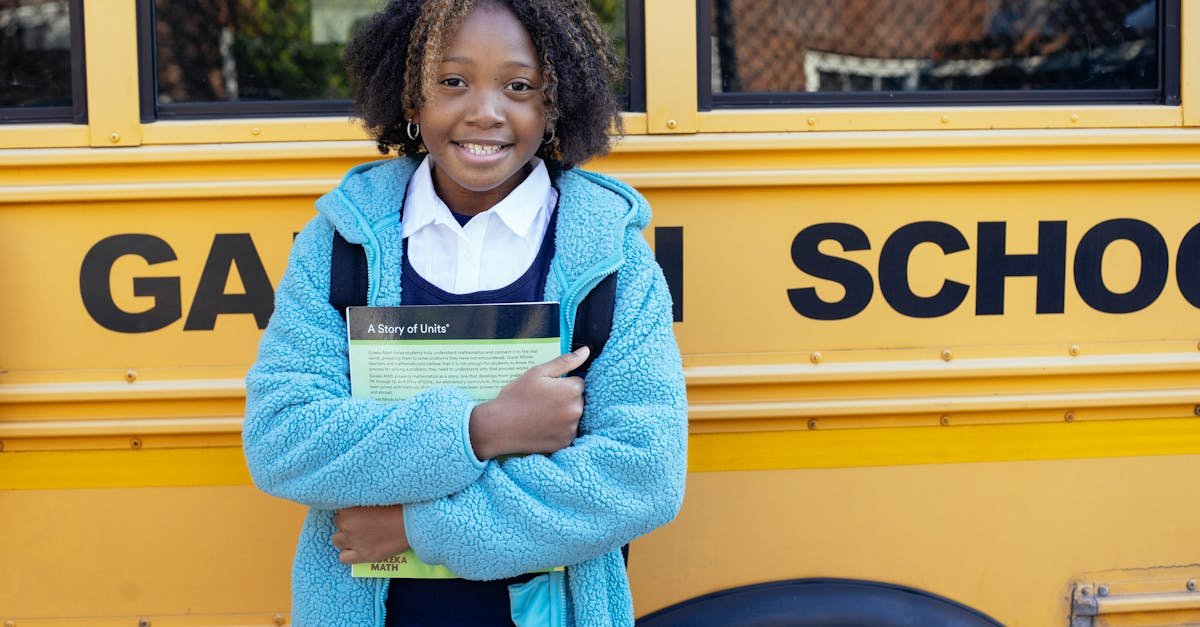 Cheerful African American girl in school uniform and warm jacket standing with textbook near yellow school bus and looking at camera