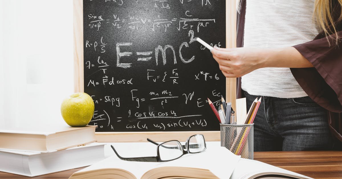 Woman writing physics equations on a blackboard with books and an apple on the desk.
