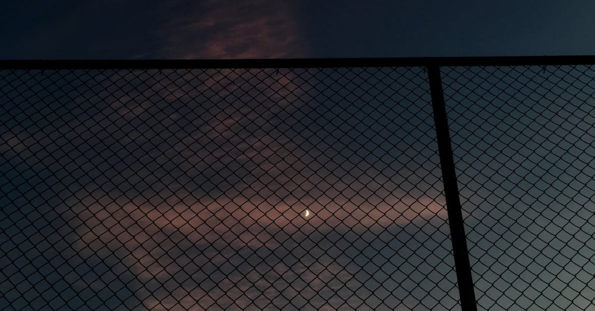 The moon peeks through a mesh wire fence against a dark sky at twilight, creating a serene silhouette.