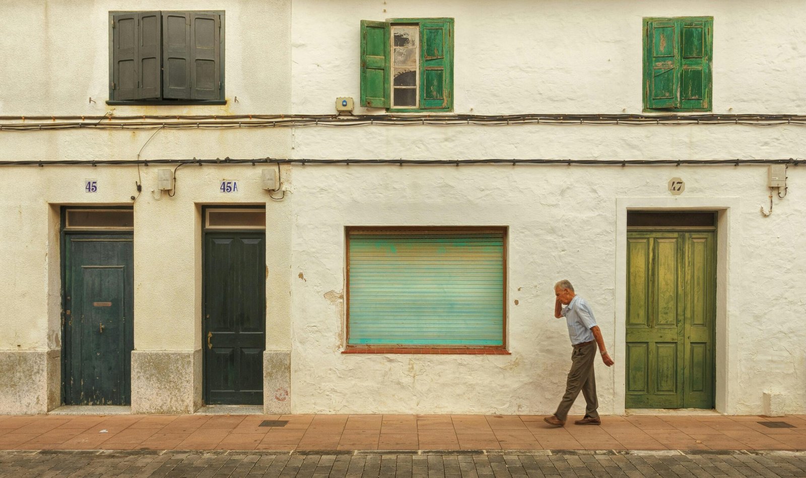 An elderly man walks past a white urban building with green doors and shutters.