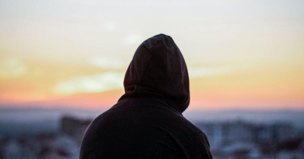 A solitary figure in a hoodie gazes at a vibrant sunset from a rooftop, creating a peaceful silhouette.
