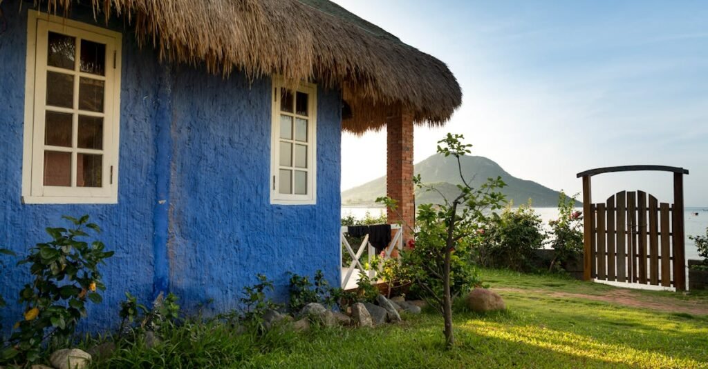 Rustic blue cottage with thatched roof by a serene lakeside and mountain view.