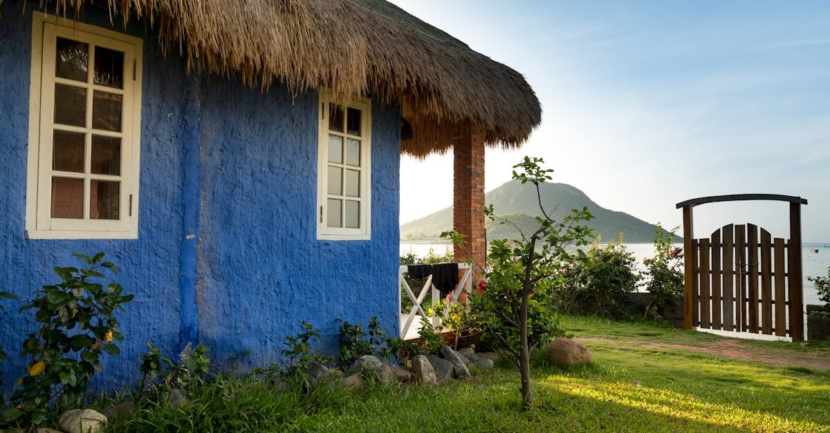 Rustic blue cottage with thatched roof by a serene lakeside and mountain view.