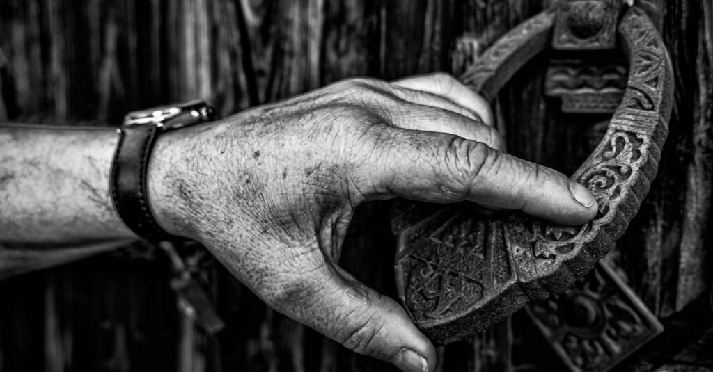 Detailed black and white photo of a hand gripping an intricately designed door knocker on a wooden gate.