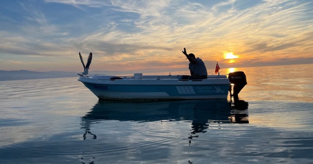 A fisherman waves from a boat during a stunning sunset over calm waters.