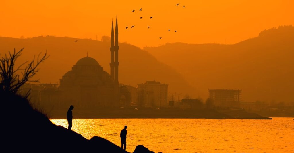 Silhouetted figures and mosque with birds at a vibrant Korgan sunset.