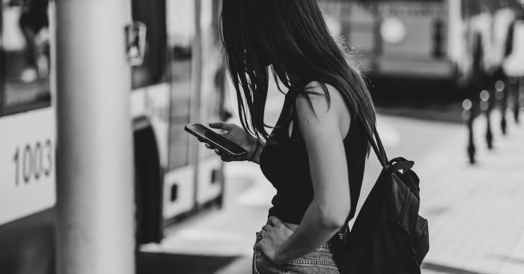 Black and white photo of a woman at a city bus stop, using her phone and wearing casual attire.