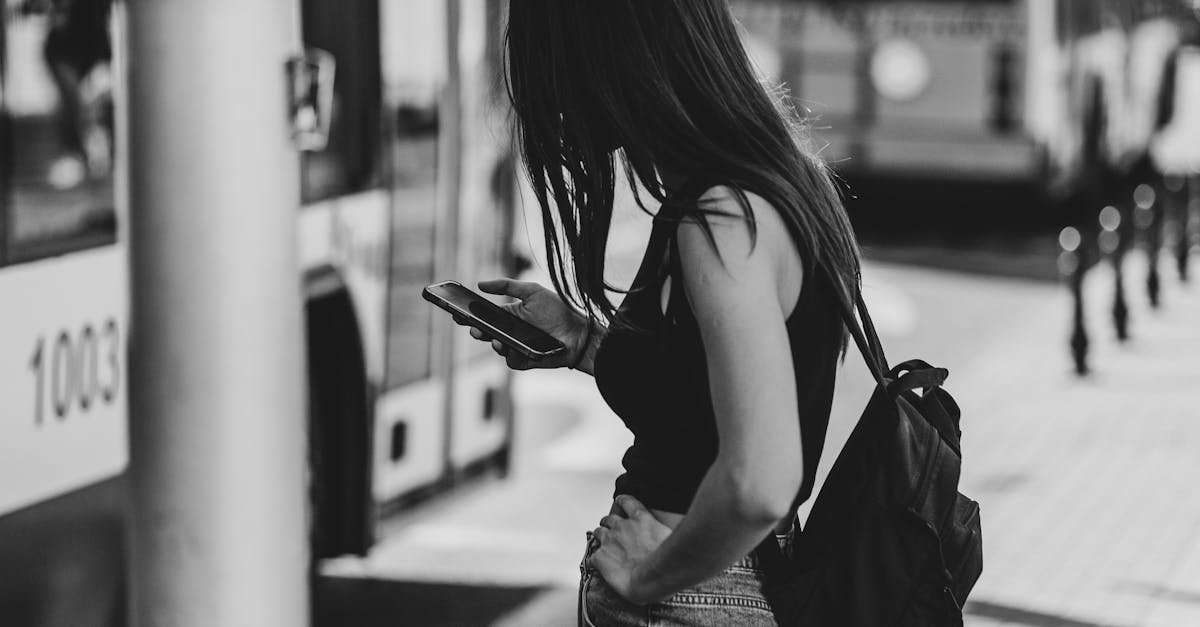 Black and white photo of a woman at a city bus stop, using her phone and wearing casual attire.