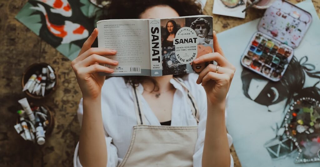 A woman lying on the floor reading a book surrounded by paintings and art supplies.
