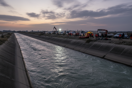 A cinematic and somber wide-angle shot of a concrete irrigation canal in Adana, Turkey, at dawn. The morning light reflects on the fast-flowing water. In the distance, rescue team vehicles and equipment are visible near the water's edge. Professional news photography style, high resolution, melancholic atmosphere.