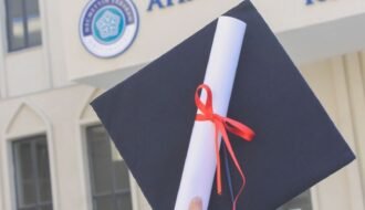 Close-up of a graduation cap and diploma held outdoors in front of a building.