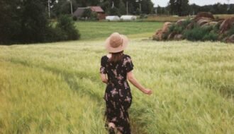 Back view of a woman in a floral dress and hat walking down a path in a lush green field.
