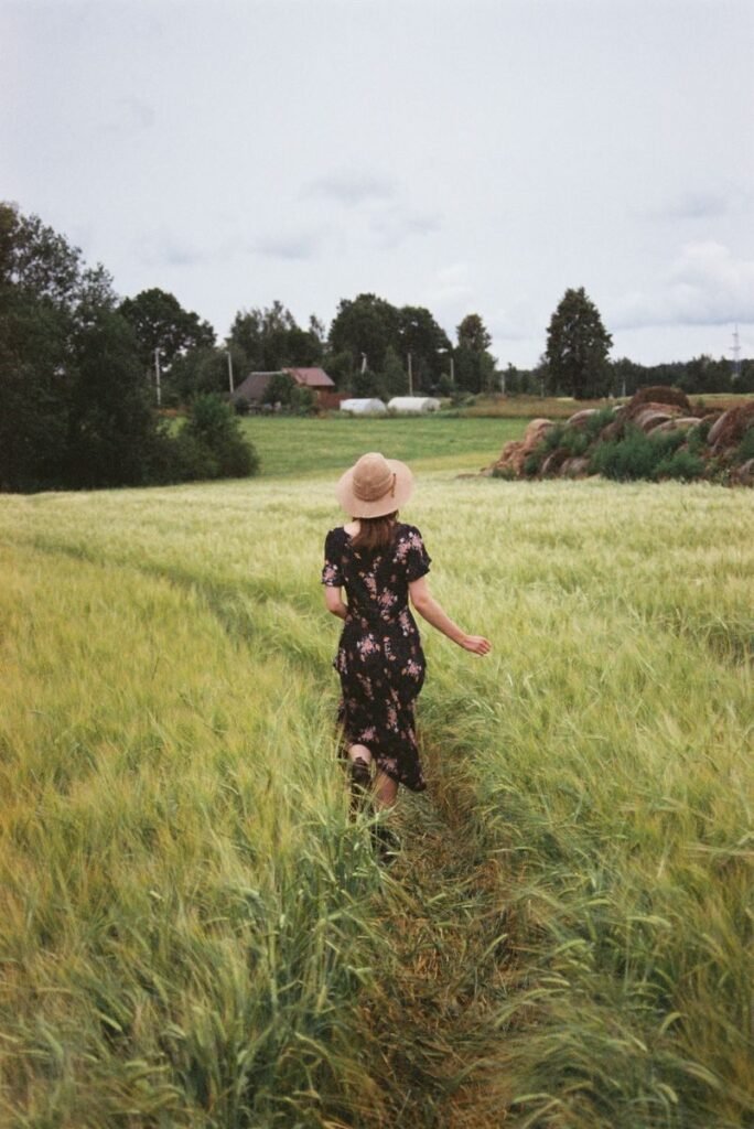 Back view of a woman in a floral dress and hat walking down a path in a lush green field.