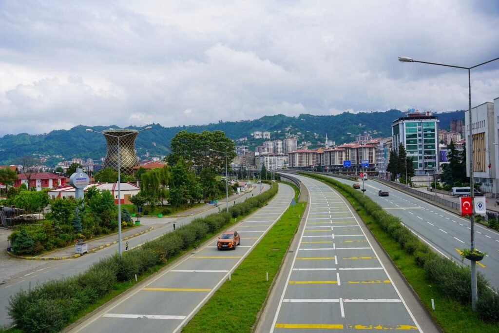 A vibrant view of a road in Rize, Türkiye, with mountains and cityscape under a cloudy sky.