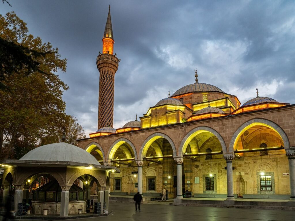 A beautiful mosque with unique architecture and a spiraled minaret, illuminated at dusk under a cloudy sky.