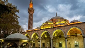 A beautiful mosque with unique architecture and a spiraled minaret, illuminated at dusk under a cloudy sky.