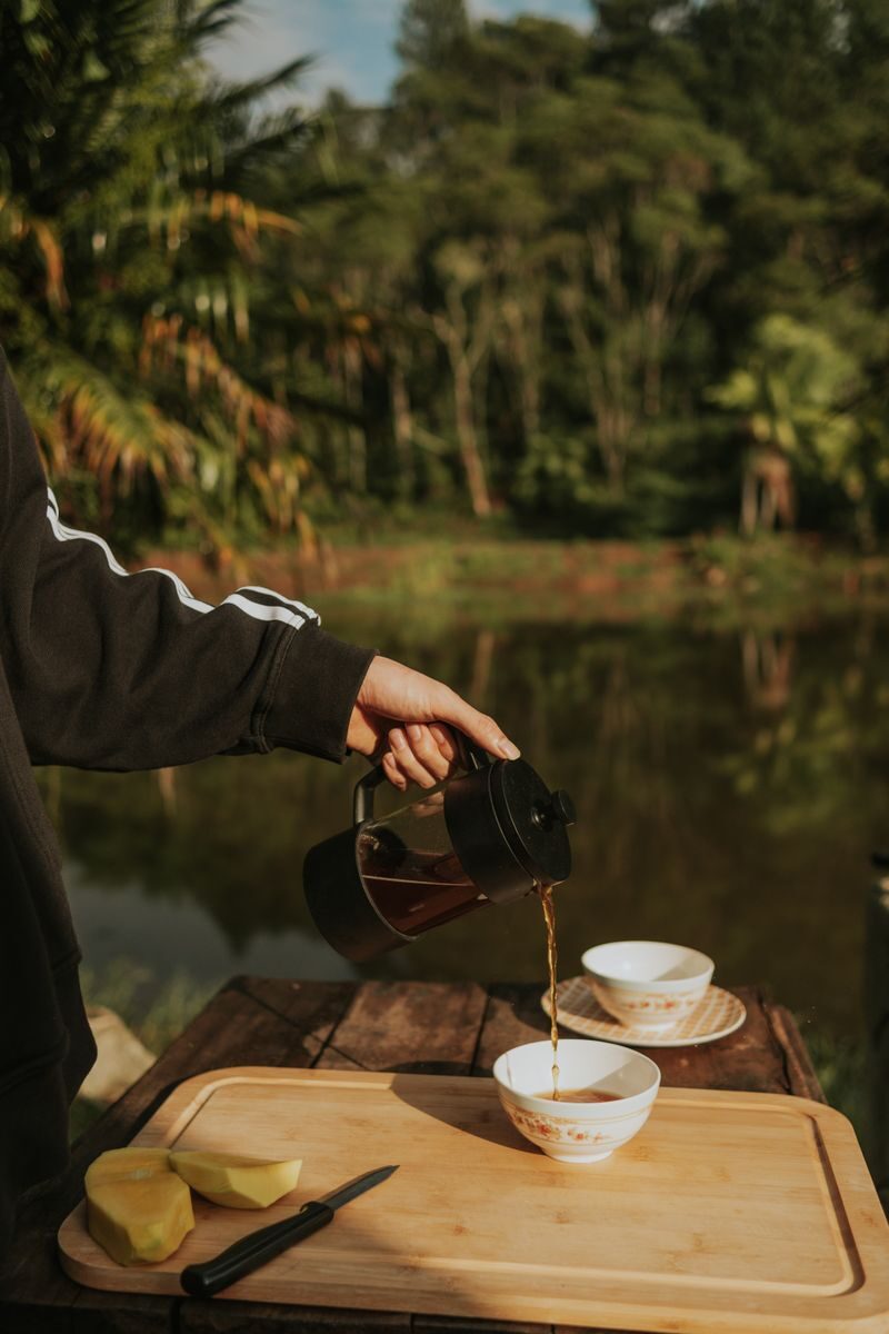Person pouring coffee from a French press by a serene pond, emphasizing outdoor relaxation.