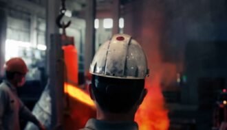 Back view of a worker in a helmet observing molten metal in an industrial setting with safety gear and smoke.